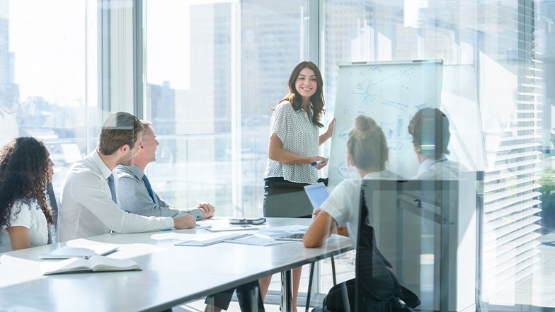 A woman stands in front of a whiteboard, presenting to a group of colleagues