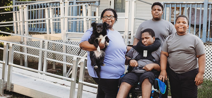 AJ is in a motorized wheelchair next to two brothers and his mom, Easter, who is holding their dog, in front of the accessible wheelchair ramp on their home.