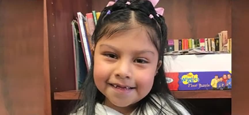 An approximately 8 year old girl with pink ribbons in her hair smiles, showing her two front teeth are missing.