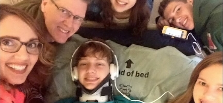 Tracy, a teen boy smiles in bed wearing a neck brace and headphones, surrounded by smiling friends and family.