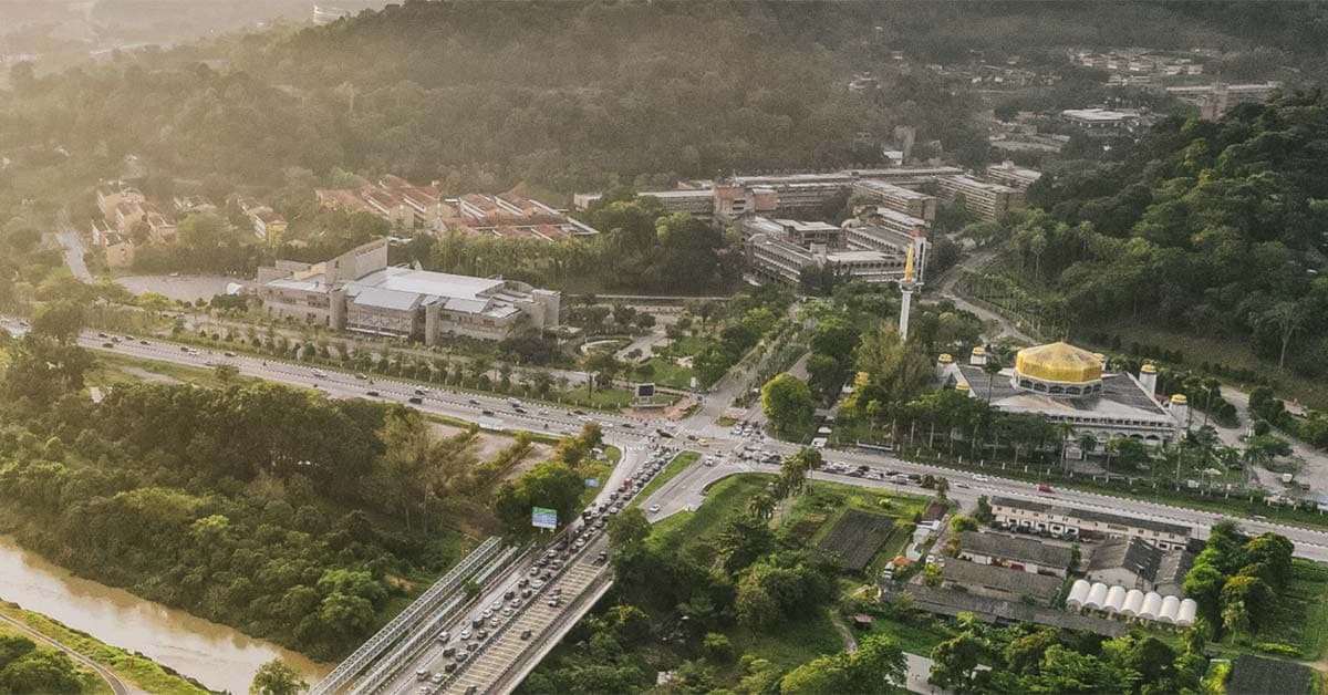 숲으로 둘러싸인 말레이시아 도시를 하늘에서 내려다본 사진. 고가도로와 금빛 돔의 모스크, 낮은 건물들이 보입니다.
